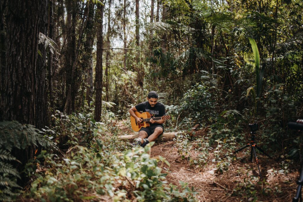 A man sits in a lush forest playing an acoustic guitar, surrounded by nature's greenery.